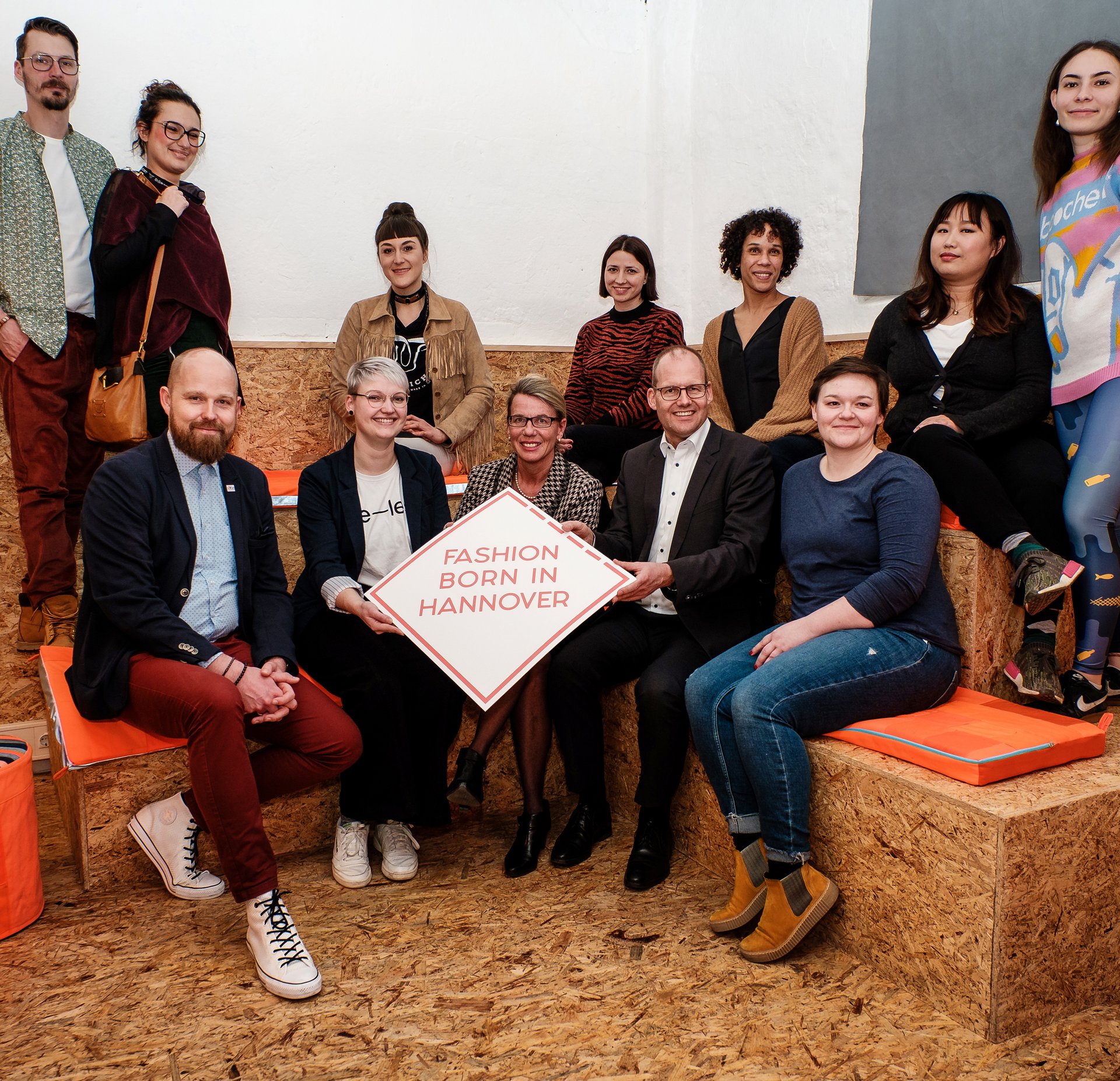 Menschen stehen und sitzen in einem Halbkreis und lächeln in die Kamera. in der Mitte halten sie ein Schild mit dem Fashion born in Hannover Logo.