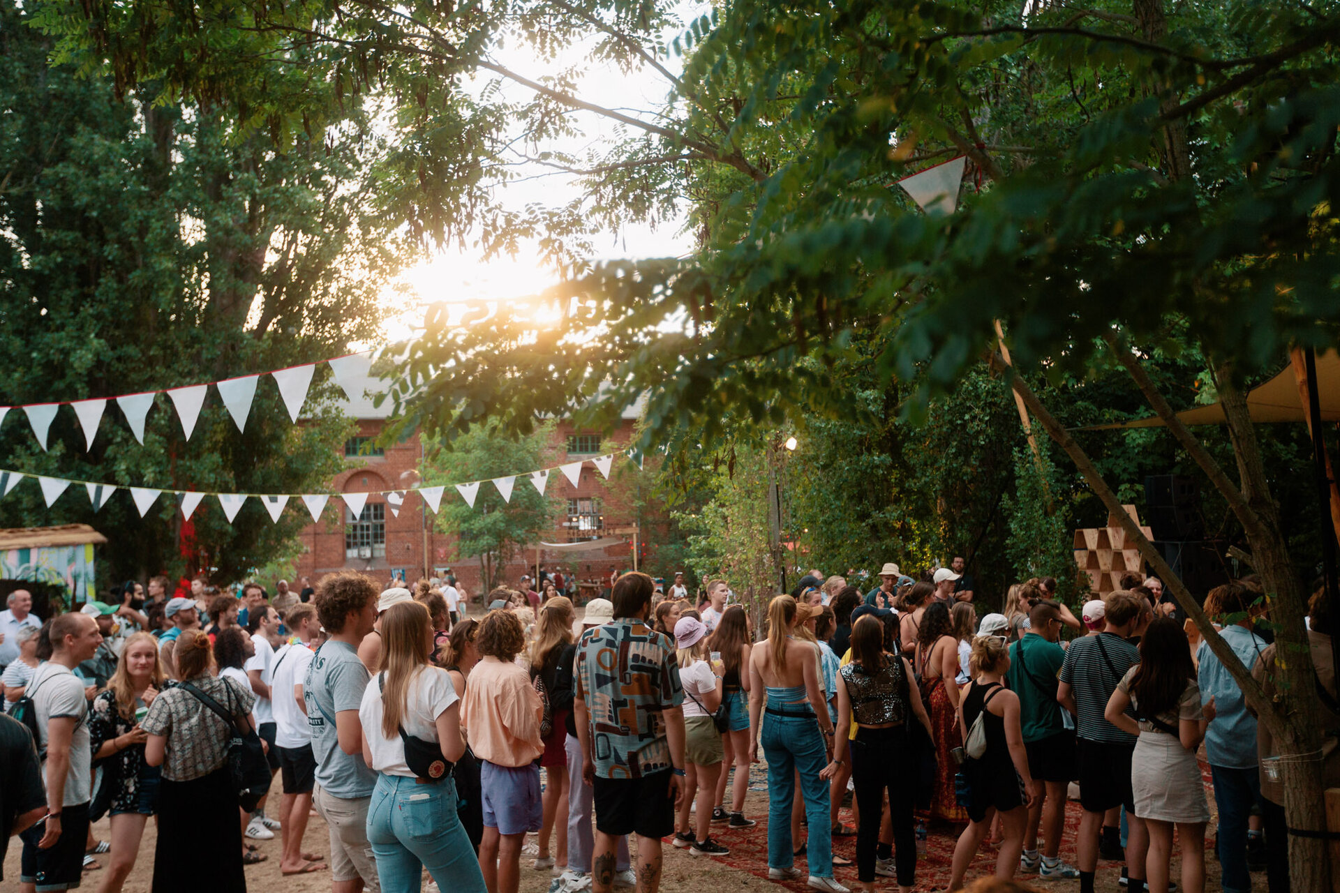 Menschen im Sommer auf einem Festival. Man sieht rechts und links Bäume, in der Mitte scheint die Sonne durch und es hängen Girlanden aus. 