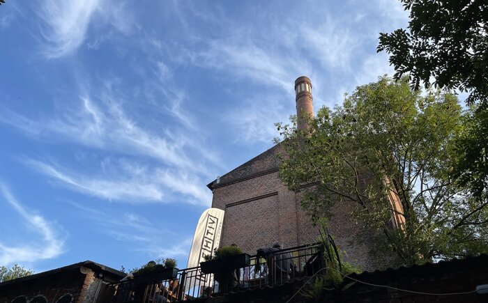 Man sieht den Gretchen Biergarten von unten fotografiert vor einem blauen Himmel. Man sieht außerdem eine kreativ-flagge.