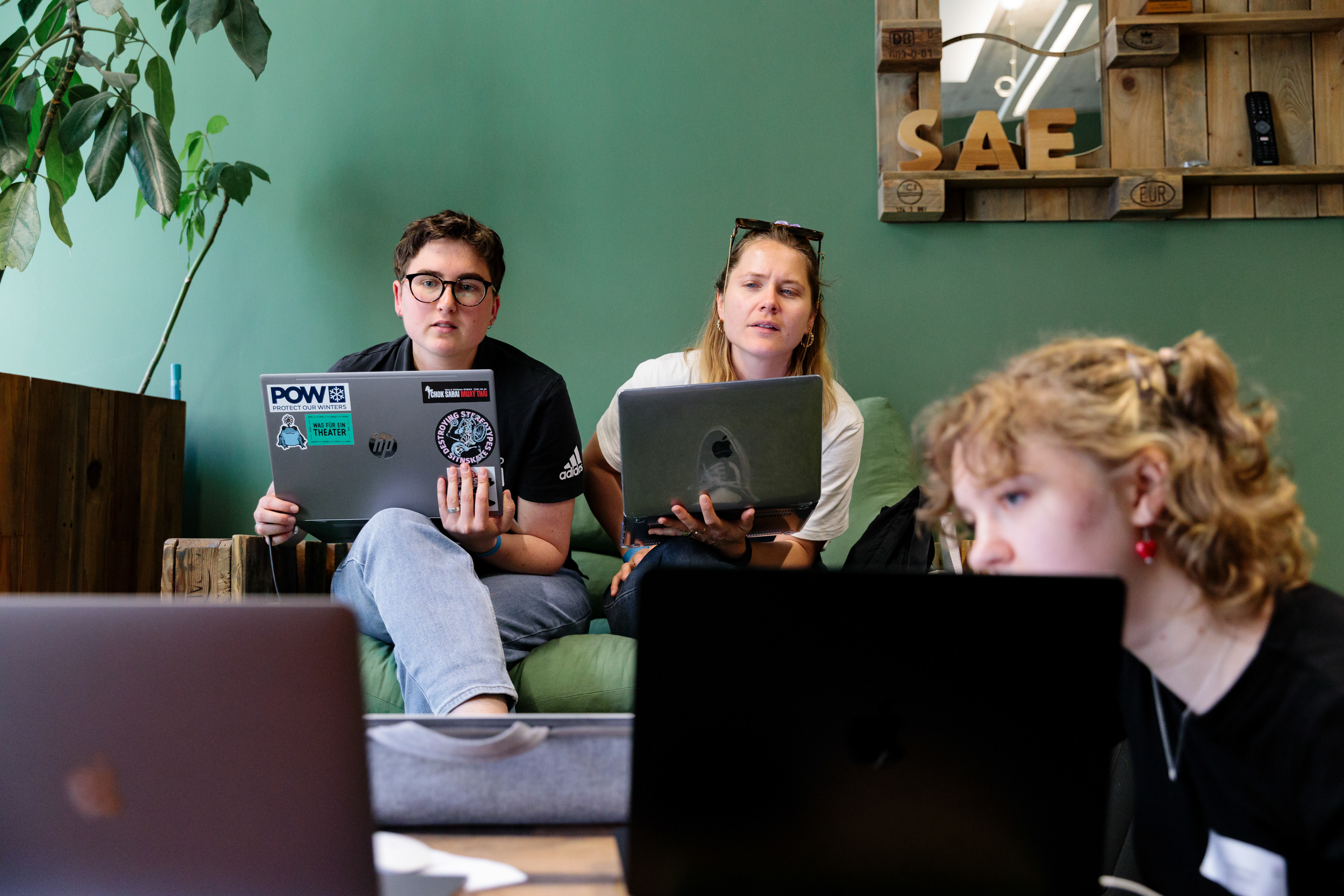 Drei Frauen mit Laptops. Eine sitzt im Vordergrund und zwei im Hintergrund auf der Couch.